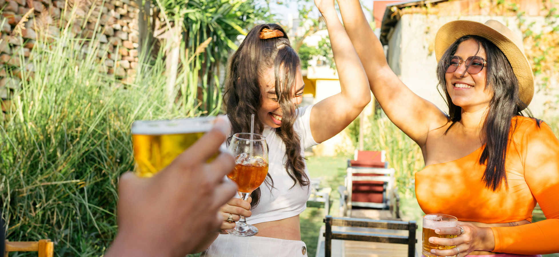 Zwei Frauen feiern in einem Garten. Eine trägt einen orangefarbenen Oberteil und eine Sonnenbrille, die andere ein weißes Top. Beide heben ihre Hände und lächeln fröhlich, während sie Getränke in den Händen halten. Im Hintergrund sind Pflanzen und Gartenmöbel zu sehen.