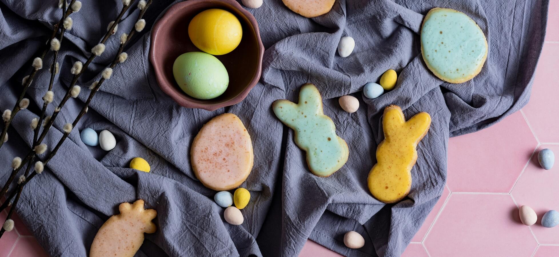 Auf einem grauen Tischtuch liegen bunte Plätzchen in Form von Hasen und Eiern. Auf der linken Seite befindet sich eine Schale mit zwei bunten Eiern. Um die Plätzchen herum sind kleine Schokoladeneier verteilt.