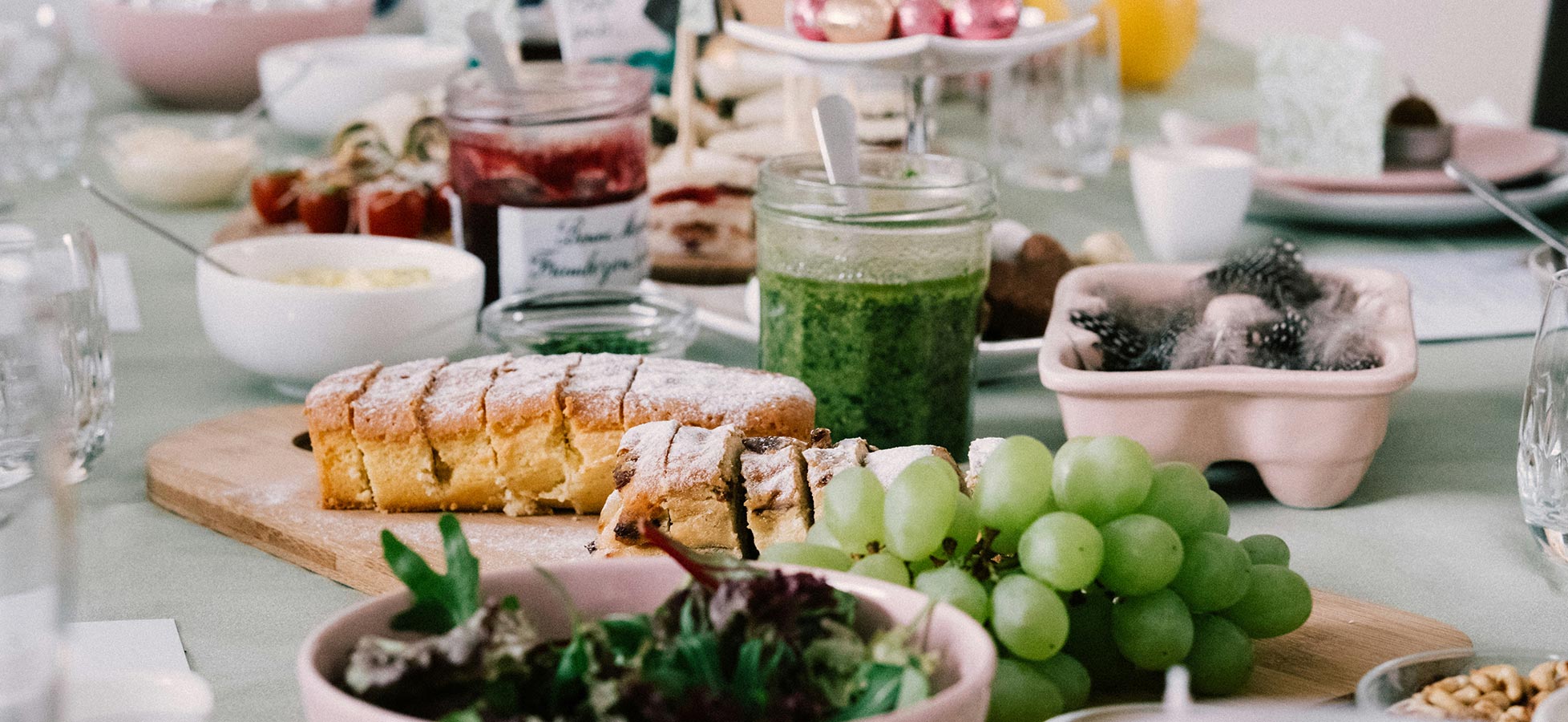 Ein festlich gedeckter Tisch mit verschiedenen Speisen: frische Trauben, ein Salat in einer Schüssel, Brot mit Puderzucker und eine grüne Soße in einem Glas. Im Hintergrund sind weitere Köstlichkeiten und Getränke auf dem Tisch verteilt.