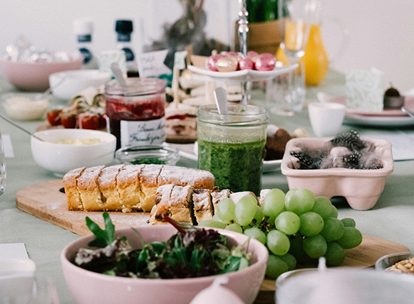 Ein festlich gedeckter Tisch mit verschiedenen Speisen: frische Trauben, ein Salat in einer Schüssel, Brot mit Puderzucker und eine grüne Soße in einem Glas. Im Hintergrund sind weitere Köstlichkeiten und Getränke auf dem Tisch verteilt.