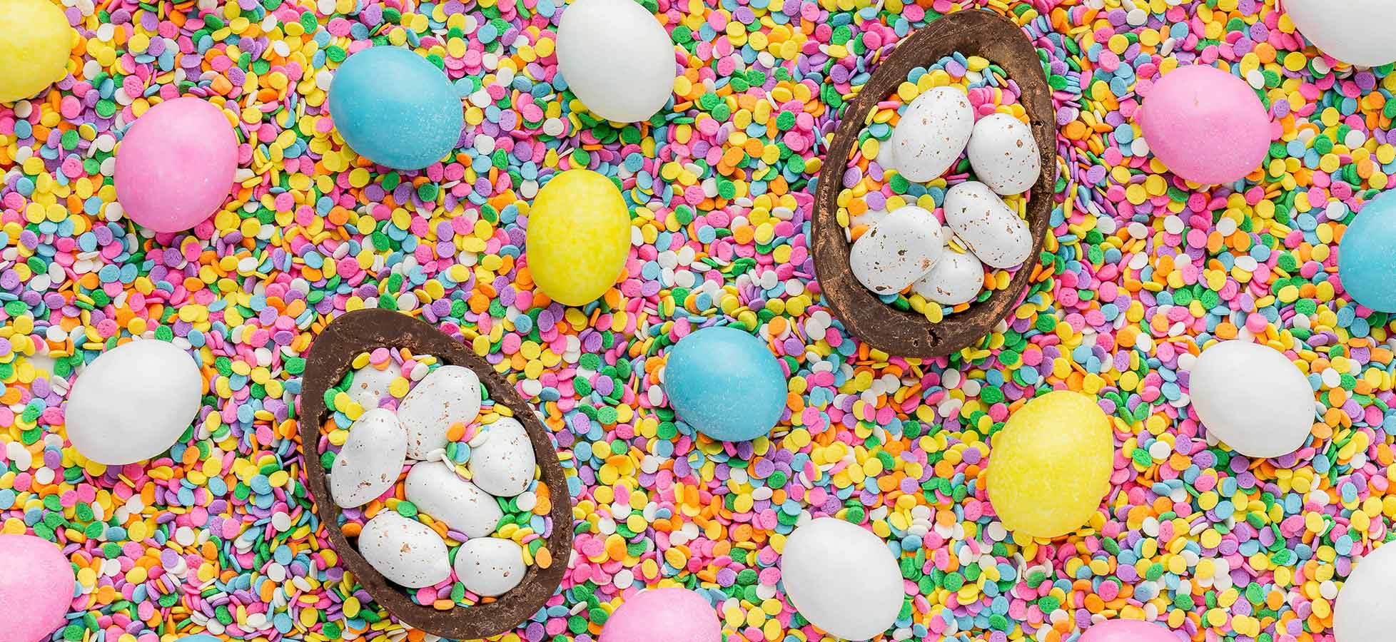 Bunte Ostereier in verschiedenen Farben (blau, gelb, pink, weiß) sind auf einem Untergrund aus bunten Zuckerkonfetti verteilt. In der Mitte befinden sich zwei Schokoladeneier, gefüllt mit kleineren weißen Schokoladeneiern. Die Szene vermittelt eine fröhliche Osterthematik.