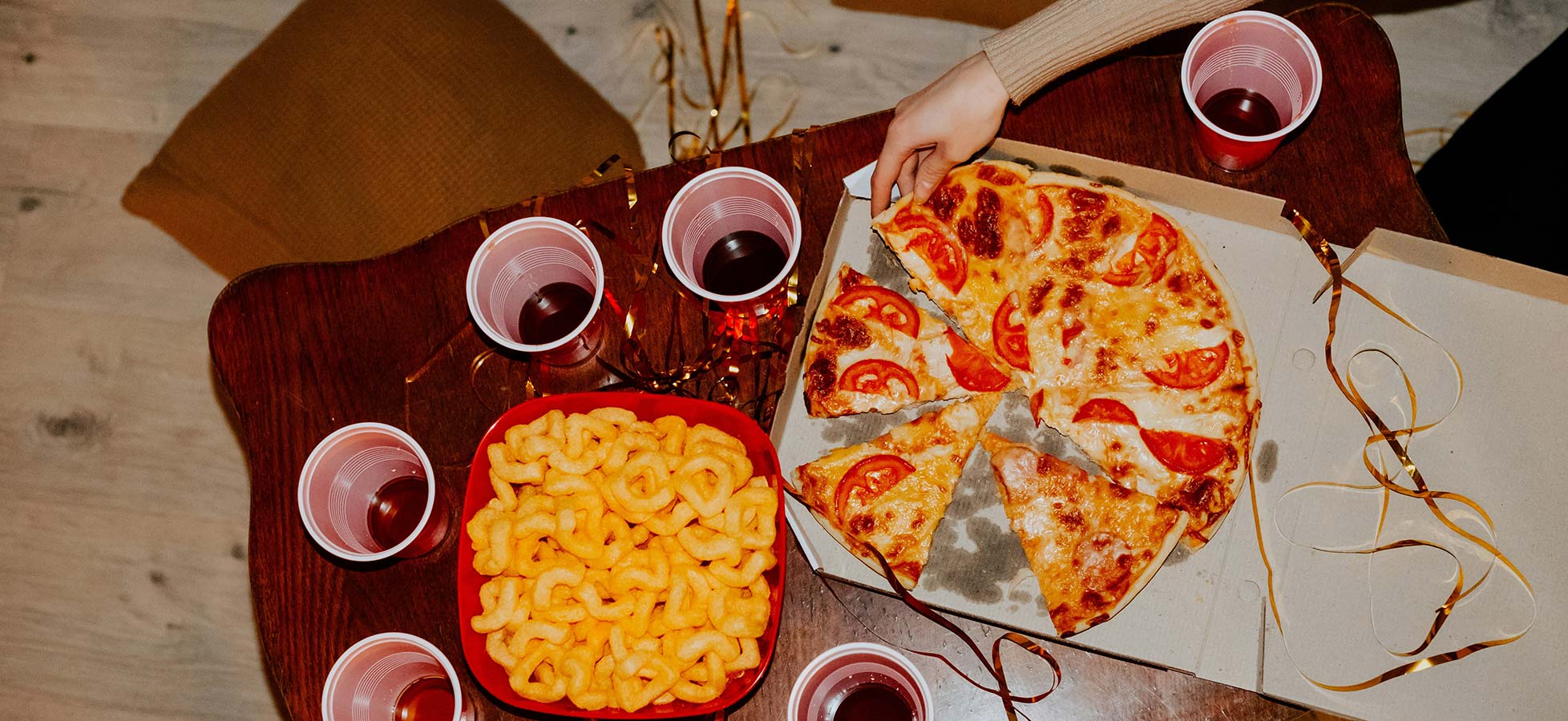 Ein Tisch ist mit einer Pizza, die in Stücke geschnitten ist, und einer Schale mit Snacks dekoriert. Rundherum stehen rote Becher mit Getränken. Es vermittelt eine gesellige Atmosphäre, ideal für eine Feier oder Zusammenkunft.