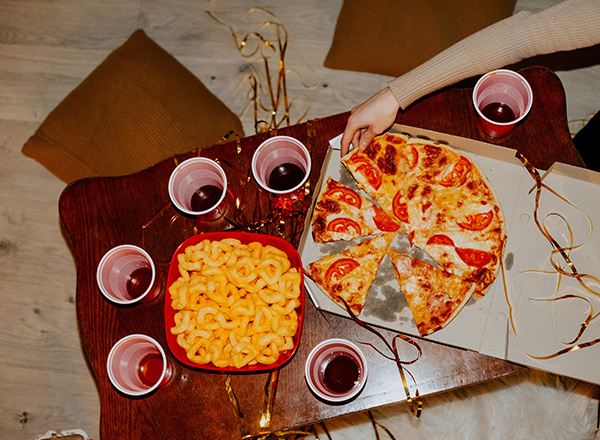 Auf einem Holztisch liegen eine große Pizza mit Tomatenscheiben, eine Schüssel mit Snacks und mehrere Einwegbecher mit Getränken. Eine Hand greift nach einem Pizzastück. Die Umgebung vermittelt eine festliche, gesellige Atmosphäre.