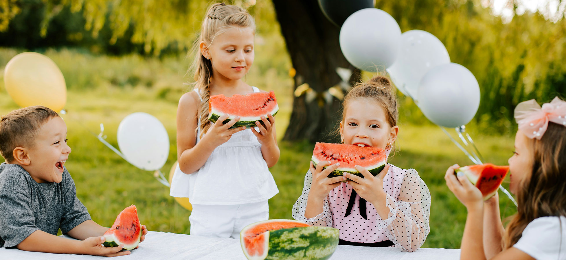 Vier Kinder sitzen an einem Tisch im Freien, halten große Stücke Wassermelone und lächeln. Sie genießen den Moment bei einem Fest, umgeben von grüner Wiese und Luftballons. Die Stimmung ist fröhlich und entspannt.