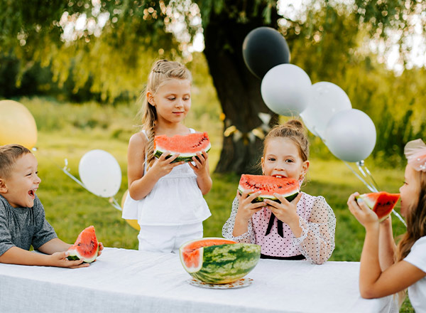 Vier Kinder sitzen an einem Tisch im Freien, halten große Stücke Wassermelone und lächeln. Sie genießen den Moment bei einem Fest, umgeben von grüner Wiese und Luftballons. Die Stimmung ist fröhlich und entspannt.