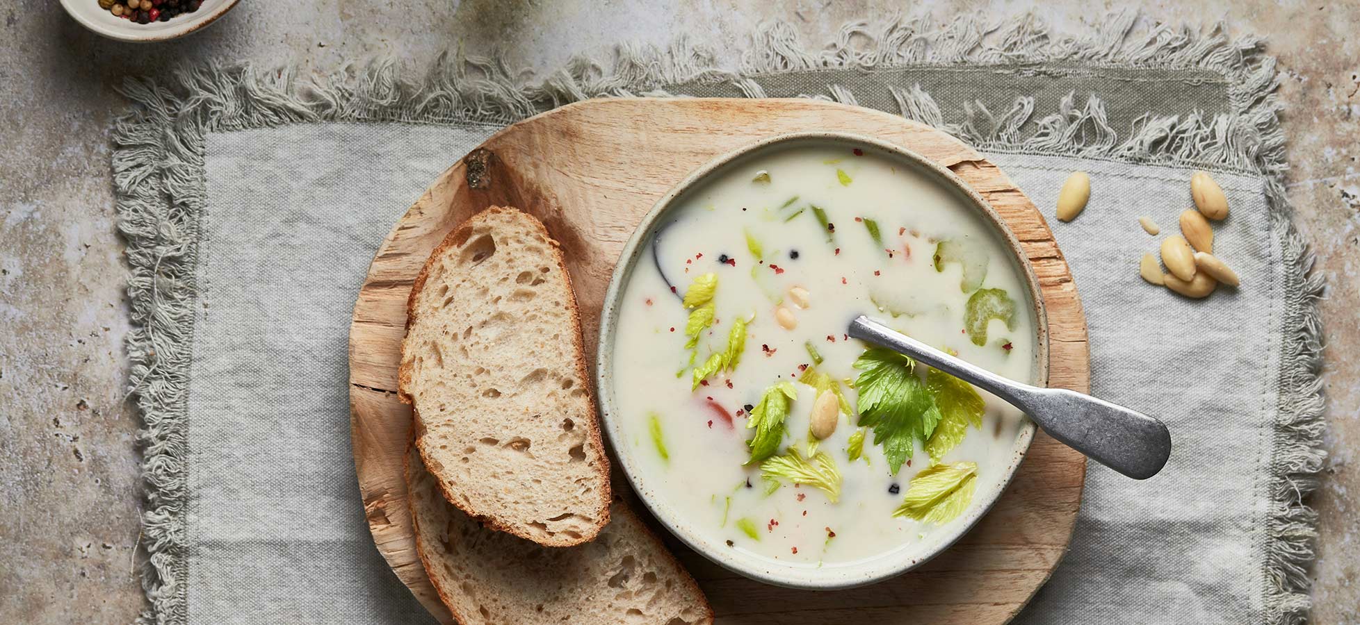Eine Schüssel cremige Suppe mit grünen Stückchen und ein paar Nüssen liegt auf einem Holzbrett. Neben der Suppe befinden sich zwei Scheiben Brot. Die Szene ist auf einem hellen Tisch mit einem Stofftuch drapiert.