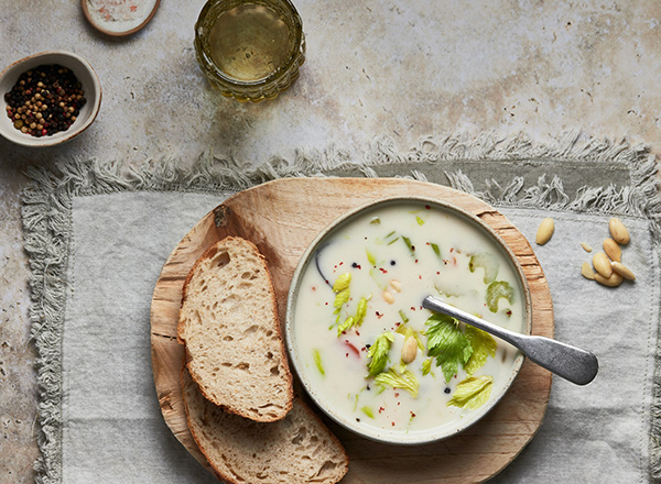 Eine Schüssel mit cremiger Suppe, garniert mit frischem Sellerie und Nüssen, steht auf einem Holzbrett. Daneben liegt ein Stück Brot. Im Hintergrund ist ein Glas und eine Schale mit Pfefferkörnern zu sehen.
