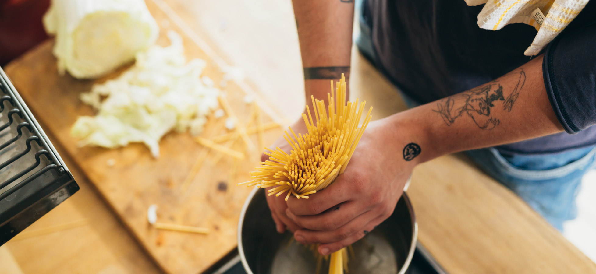 Eine Person hält eine Portion ungekochte Spaghetti über einen Topf mit Wasser. Im Hintergrund ist ein Schneidebrett mit vorbereiteten Zutaten zu sehen. Es herrscht eine gemütliche Kochatmosphäre.