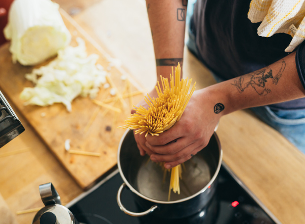 Eine Person hält eine Handvoll Spaghetti über einen Topf mit kochendem Wasser, bereit zum Kochen. Im Hintergrund sind ein Holzbrett mit Kohlresten und eine Herdplatte zu sehen.