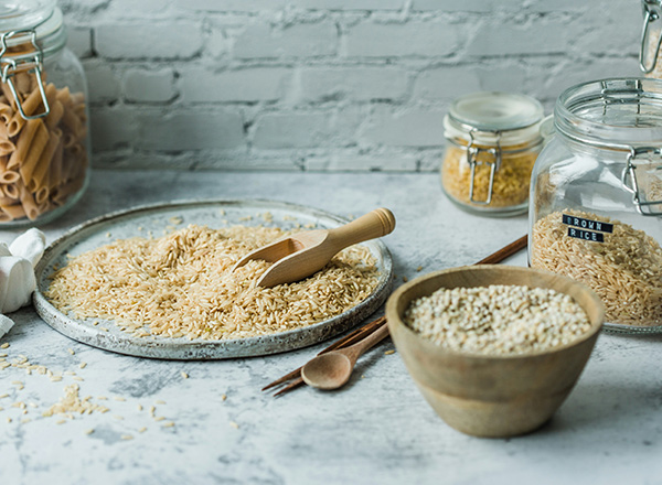 Eine flache Schale mit braunem Reis und einem Holzlöffel ist im Vordergrund. Daneben stehen zwei Schalen mit verschiedenen Reissorten. Im Hintergrund sind mehrere Gläser mit trockenen Lebensmitteln. Der Tisch hat eine helle, strukturierte Oberfläche.