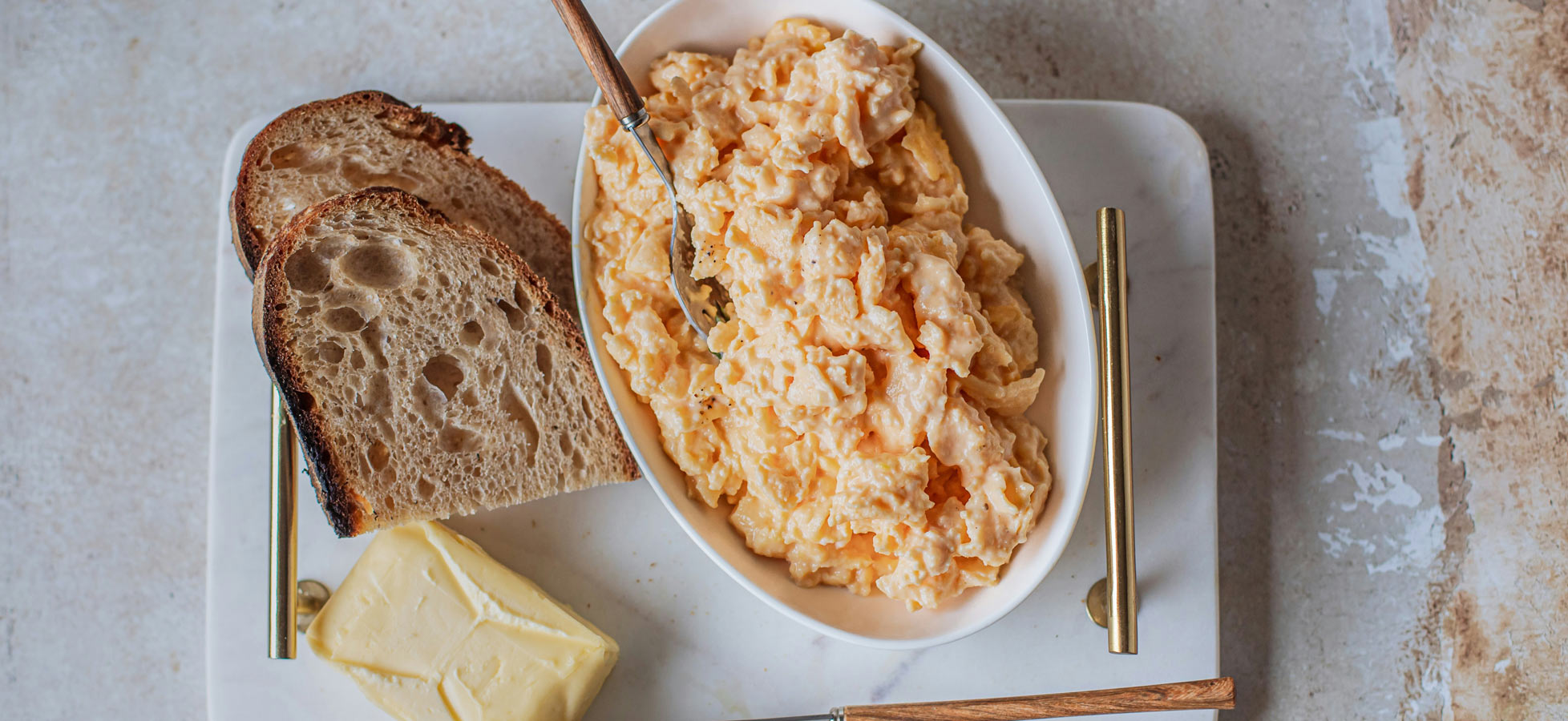 Ein Teller mit Rührei und einer Gabel, daneben liegen zwei Scheiben Brot und ein Stück Butter. Die Speisen sind auf einer weißen Servierplatte angerichtet.