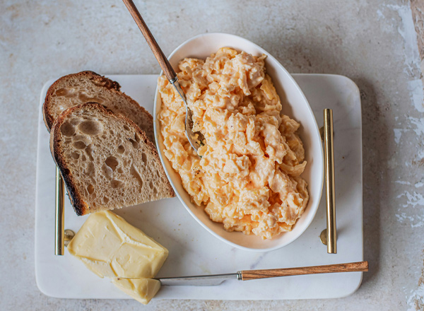 Ein weißes Schälchen mit Rührei befindet sich auf einer Platte. Daneben liegen zwei Scheiben Brot und ein Stück Butter. Besteck mit Holzgriffen ist ebenfalls vorhanden, was auf eine gemütliche Mahlzeit hindeutet.