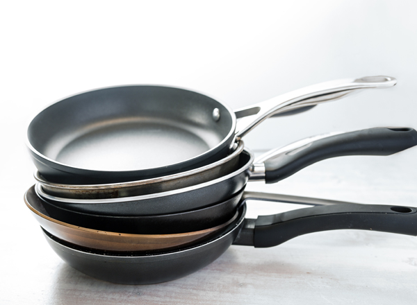 Stapel von Pfannen in verschiedenen Größen und Materialien. Die oberste Pfanne ist schlicht schwarz, darunter befinden sich eine Pfanne mit silbernem Rand und eine mit kupferfarbener Unterseite. Alle haben lange, ergonomische Griffe.