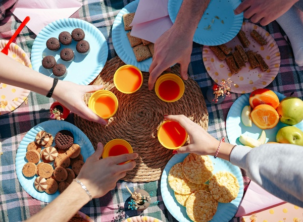 Mehrere Hände halten orangefarbene Getränke, während sie sich um einen Picknicktisch versammeln. Auf bunten Tellern liegen verschiedene Snacks, darunter Kekse, Obst und Cracker. Die Atmosphäre ist lebhaft und einladend, mit einer Decke im Hintergrund.