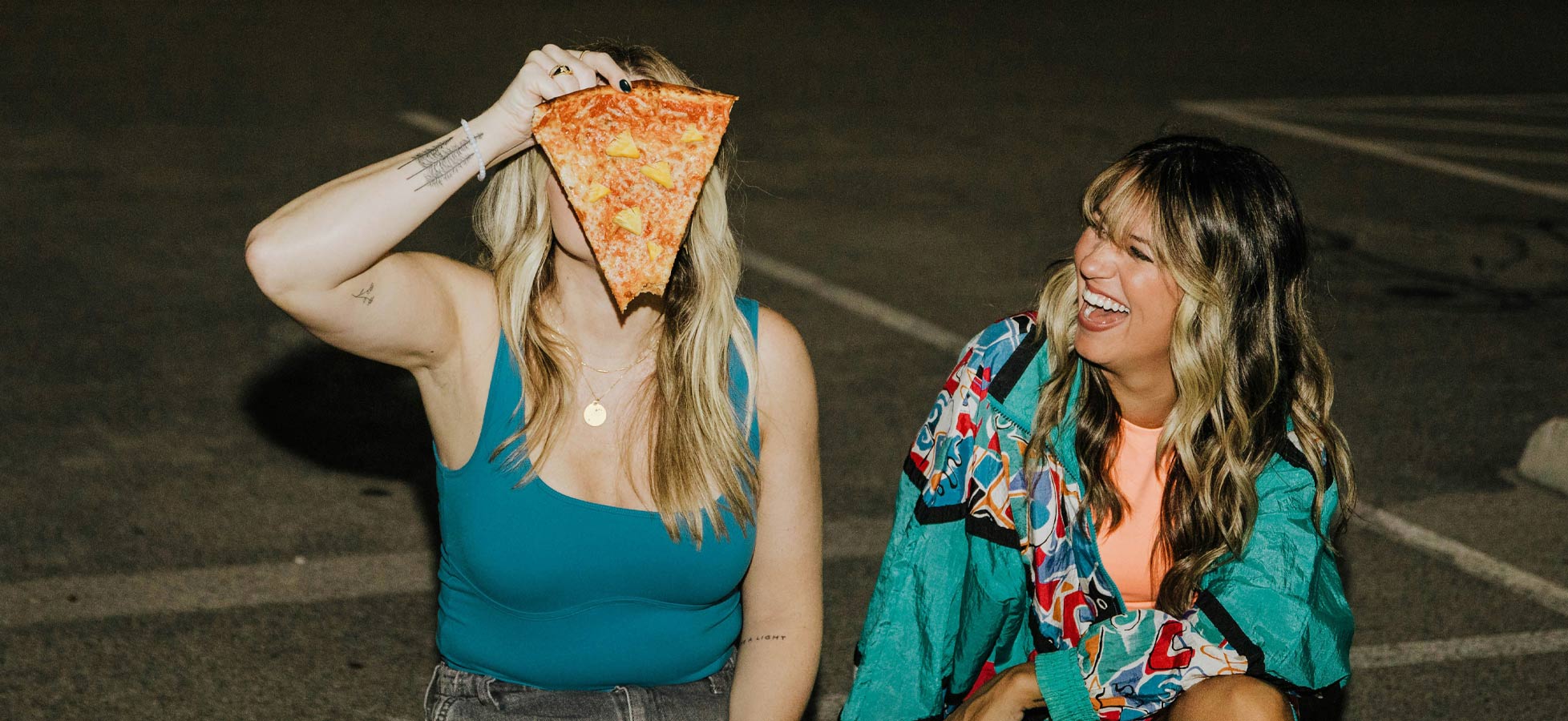 Zwei Frauen sitzen auf einem Parkplatz. Eine Frau hält ein großes Stück Pizza vor ihr Gesicht, während die andere fröhlich lacht. Beide wirken entspannt und haben Spaß miteinander. Die Umgebung ist dunkel und wird nur schwach beleuchtet.