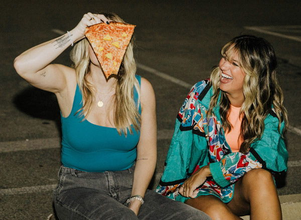 Zwei Frauen sitzen in einem Parkplatz. Eine Frau hält ein großes Stück Pizza vor ihr Gesicht, während die andere Frau lachend daneben sitzt. Beide wirken entspannt und genießen den Moment.