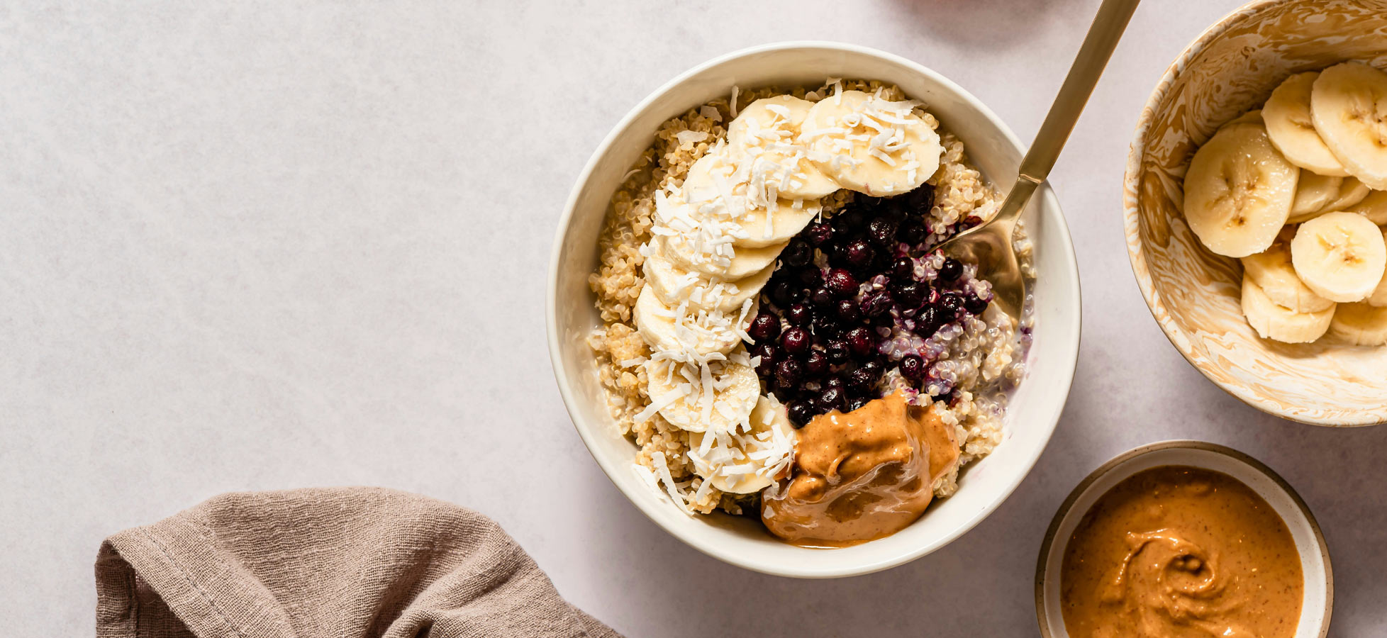 Eine Schüssel Haferbrei mit Bananenscheiben, Heidelbeeren und Erdnussbutter. Neben der Schüssel befinden sich eine kleine Schale mit weiteren Bananenscheiben. Der Hintergrund ist hell und schlicht.
