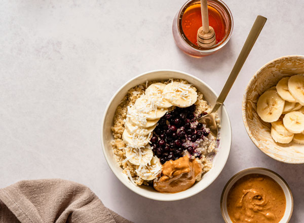 Eine Schüssel Haferbrei mit Bananenscheiben, Heidelbeeren und Erdnussbutter. Neben der Schüssel befinden sich eine kleine Schale mit weiteren Bananenscheiben und ein Glas Honig mit einem Holzlöffel. Der Hintergrund ist hell und schlicht.