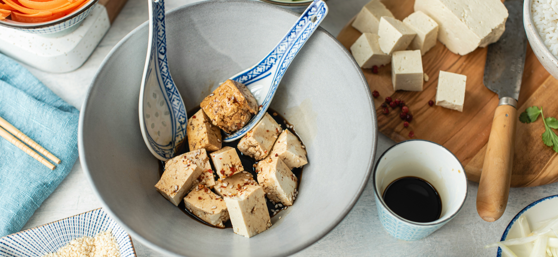 Eine große Schüssel enthält gewürzten Tofu, der in Stücke geschnitten ist. Zwei Löffel liegen in der Schüssel. Daneben sind weitere Tofu-Würfel sowie kleinere Schüsseln mit Sojasauce und anderen Zutaten zu sehen.