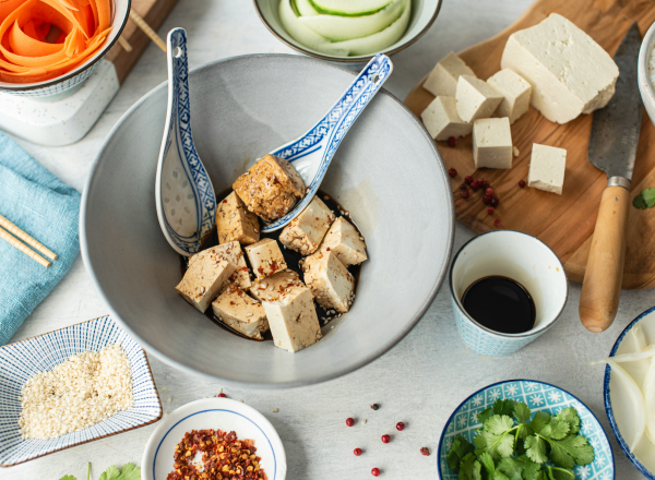 Eine große Schüssel mit gewürfeltem Tofu in Dunkelsoße, garniert mit Sesamsamen, steht im Mittelpunkt. Rundherum sind verschiedene Zutaten wie Karotten, Gurken, frischer Koriander, Salz, Chili und Sojasoße in kleinen Schalen angeordnet. Das Bild vermittelt eine bunte, gesunde Essenszubereitung.
