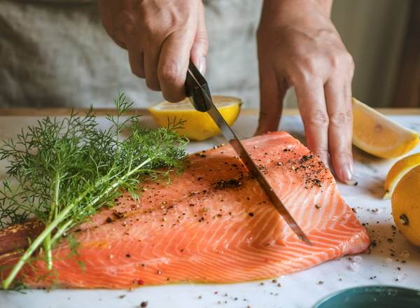Eine Person schneidet mit einem Messer ein Stück Lachs auf einem Holzbrett. Um den Fisch herum liegen frische Kräuter und gefüllte Zitronenhälften. Der Lachs ist gewürzt mit Pfeffer.