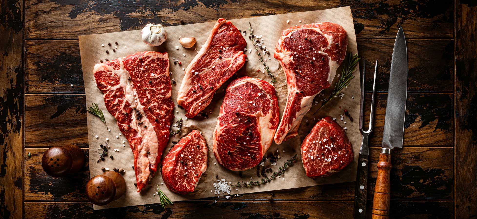 Eine Holzplatte zeigt mehrere verschiedene rohe Fleischstücke, darunter Steaks mit intensivem Marmorierungsmuster. Einige Gewürze, wie Salz und Pfeffer, sind verstreut, sowie frische Kräuter. Rechts sind ein großes Messer und eine Gabel mit Holzgriffen zu sehen.