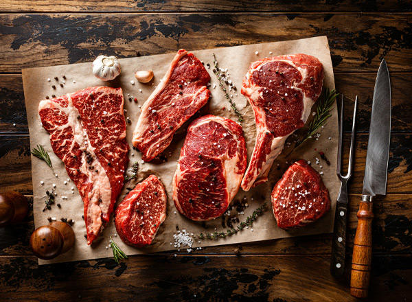 Auf einem Holzbrett liegen verschiedene Stücke rohes Fleisch, darunter entbeinte Steaks und Fleischstücke mit Fettanteilen. Um das Fleisch sind Gewürze, Knoblauchzehen und Kräuter arrangiert. Neben dem Brett liegen ein scharfes Messer und ein Kräutermesser.