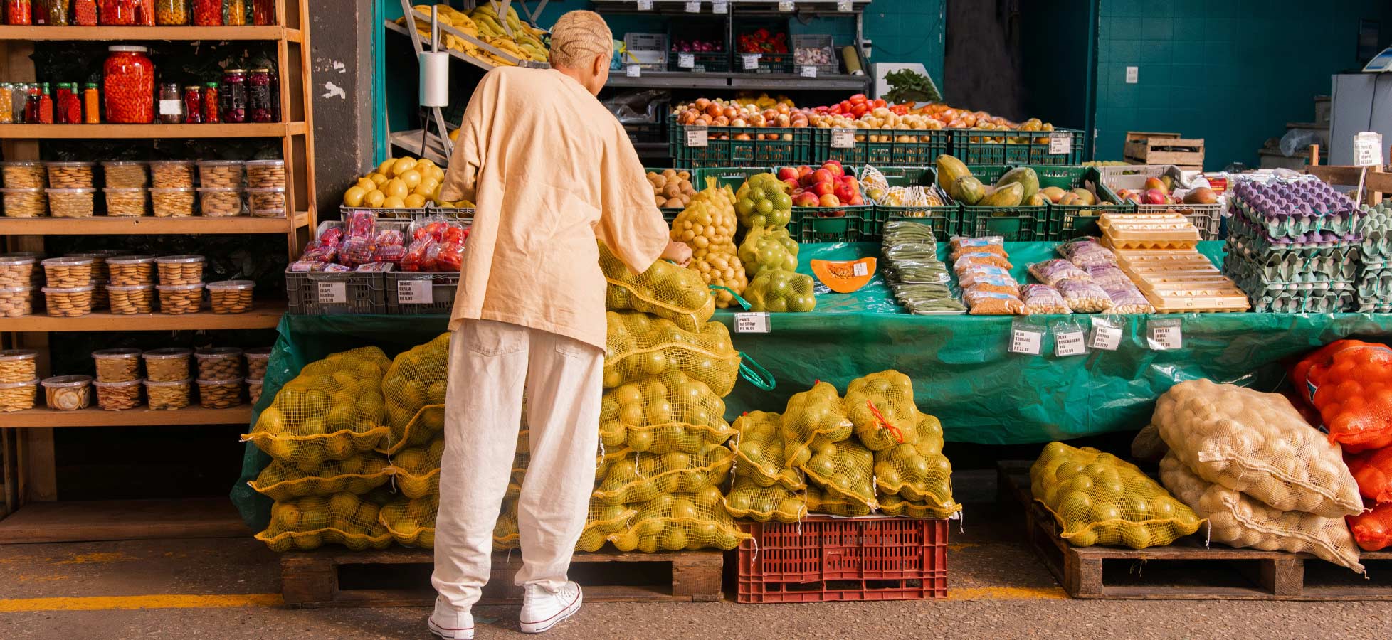 Eine Person steht vor einem Marktstand und betrachtet Menge an Obst und Gemüse, darunter Äpfel, Bananen und verschiedene Sorten Zitrusfrüchte. Der Stand ist voll mit Obst in Netzen und Kisten, während Vorratsbehälter im Hintergrund sichtbar sind.