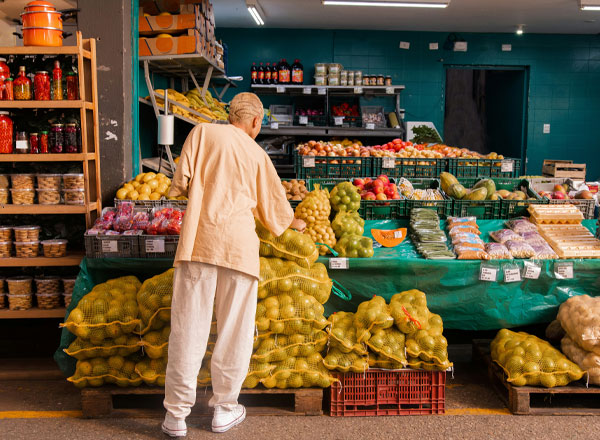 Eine Person steht vor einem Marktstand und betrachtet Menge an Obst und Gemüse, darunter Äpfel, Bananen und verschiedene Sorten Zitrusfrüchte. Der Stand ist voll mit Obst in Netzen und Kisten, während Vorratsbehälter im Hintergrund sichtbar sind.