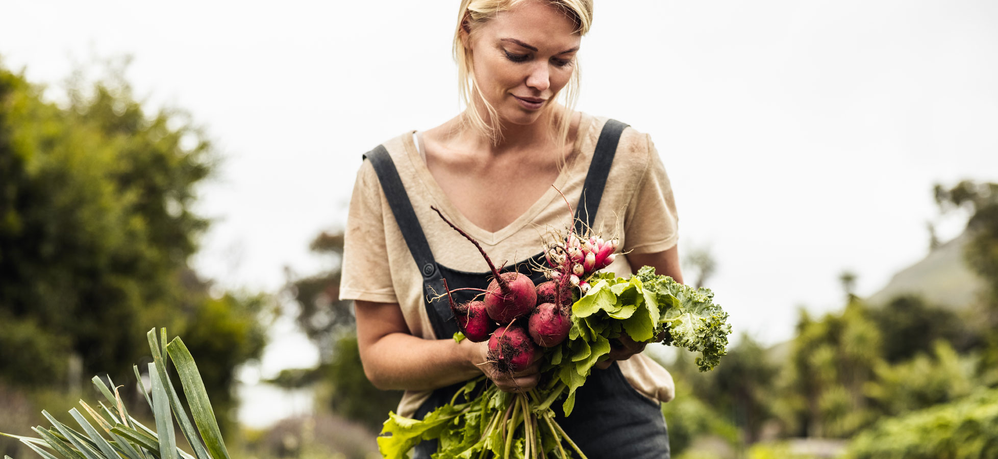 Eine Frau steht in einem Garten und hält frisches Gemüse in den Händen, darunter Radieschen und Blätter. Sie trägt ein einfaches T-Shirt und einen Latz, während sie aufmerksam auf die Ernte schaut. Im Hintergrund sind grüne Pflanzen und eine bewölkte Himmel sichtbar.