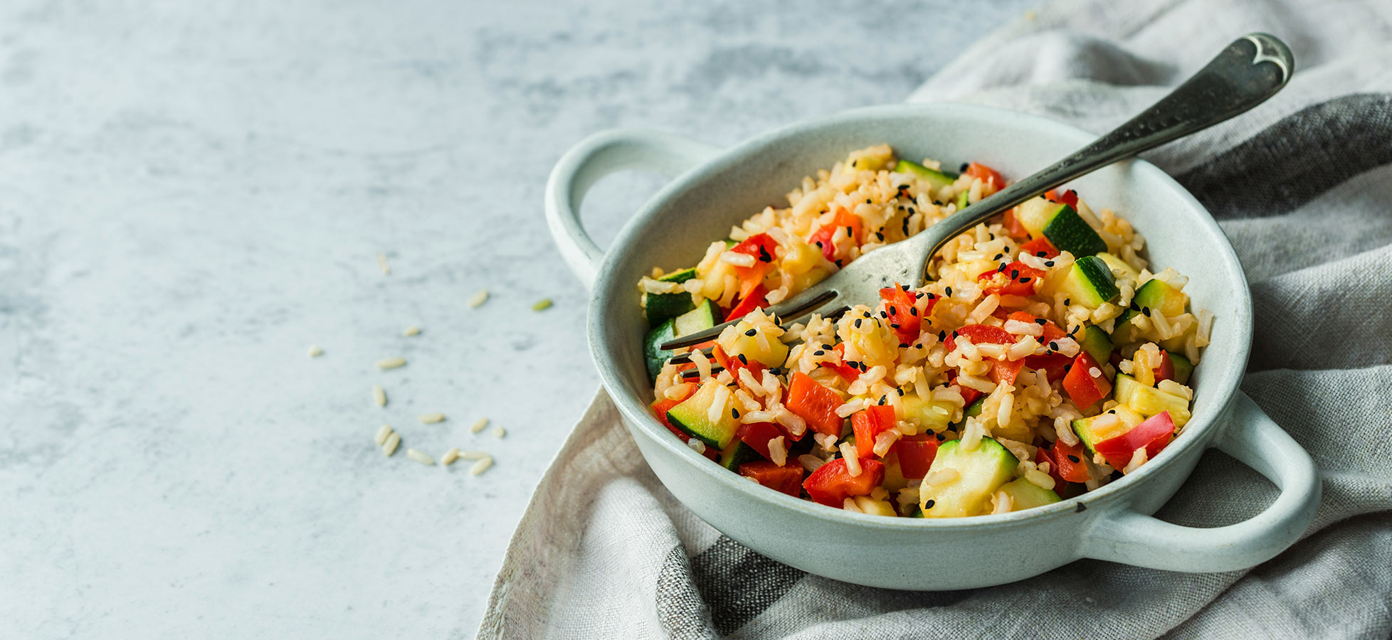 Schüssel mit buntem Gemüse-Reis, bestehend aus verschiedenen Gemüsesorten wie Paprika und Zucchini, garniert mit einer Gabel. Die Schüssel steht auf einer Stoffserviette auf einem hellen Tisch.