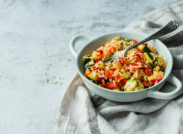 Eine Schüssel mit buntem Gemüsereis, der Zucchini, Paprika und anderen Gemüsesorten enthält. Ein Löffel ist im Gericht platziert. Der Hintergrund ist neutral und hell.