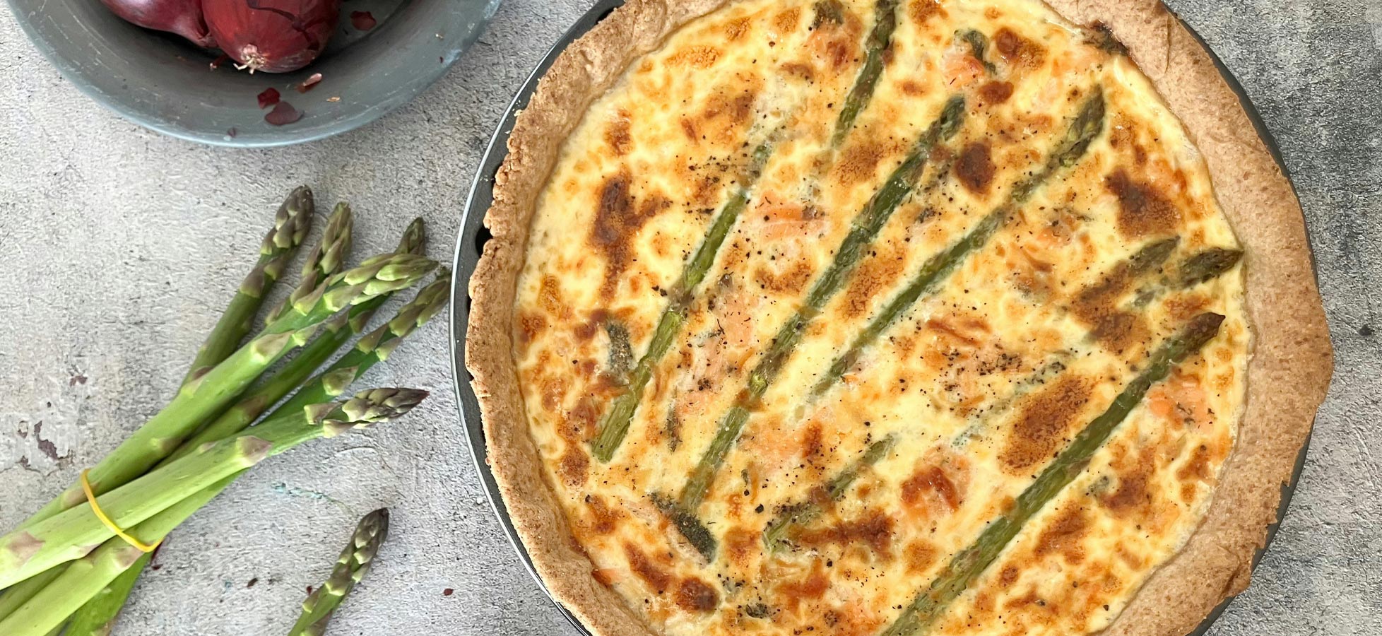 Ein frisch gebackener Quiche mit grünem Spargel ist im Vordergrund zu sehen. Daneben liegen einige grüne Spargelstangen und rote Zwiebeln in einer Schale. Die Quiche hat eine goldbraune Kruste und ist mit sparsamen Streifen von Spargel dekoriert.