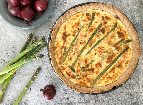 Ein frisch gebackener Quiche mit grünem Spargel ist im Vordergrund zu sehen. Daneben liegen einige grüne Spargelstangen und rote Zwiebeln in einer Schale. Die Quiche hat eine goldbraune Kruste und ist mit sparsamen Streifen von Spargel dekoriert.