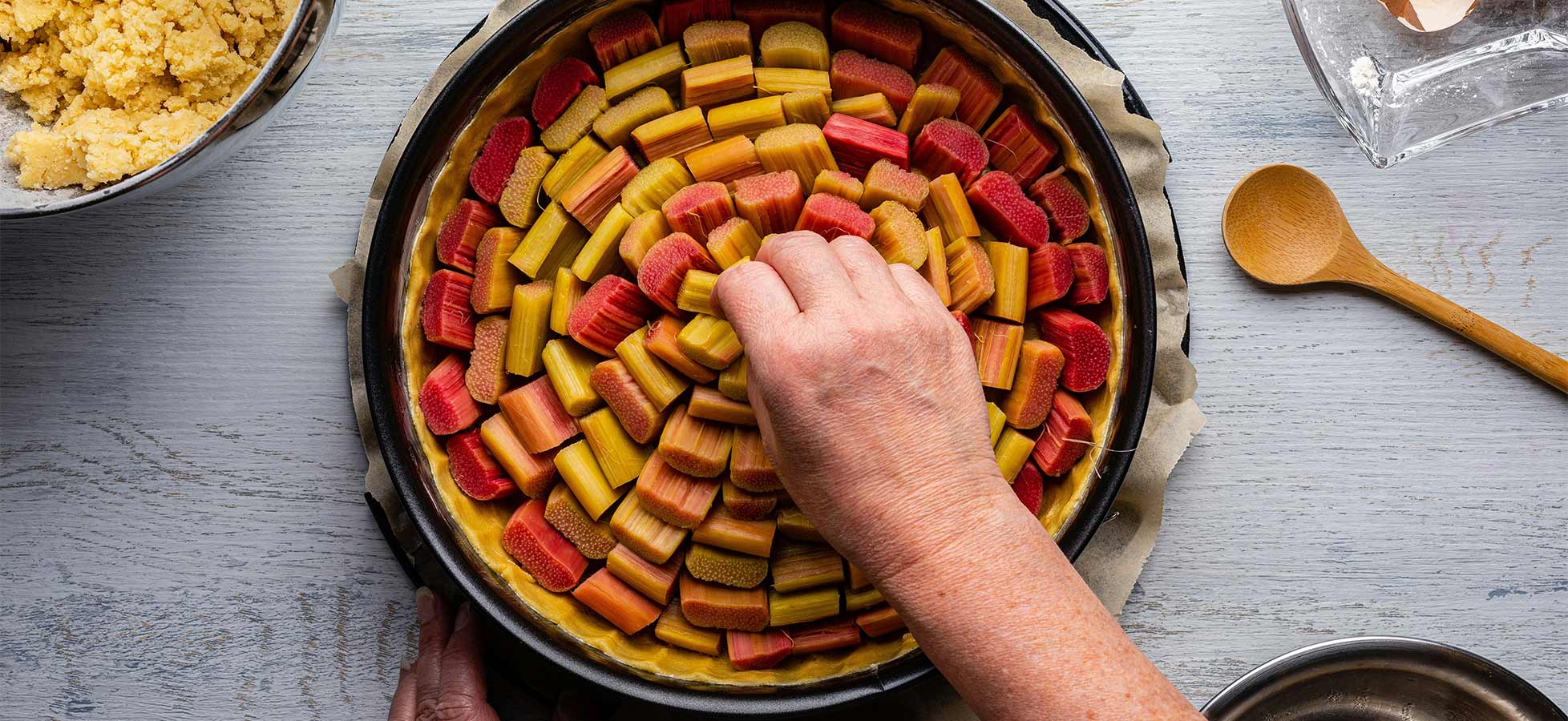 Eine Hand platziert bunte Rhabarberstücke in einem runden Tortenblech. Die Rhabarberstücke sind gleichmäßig angeordnet und zeigen eine Kombination aus grünen und roten Farben. Im Hintergrund sind weitere Zutaten in Schalen sichtbar, aber nicht im Fokus der Darstellung.