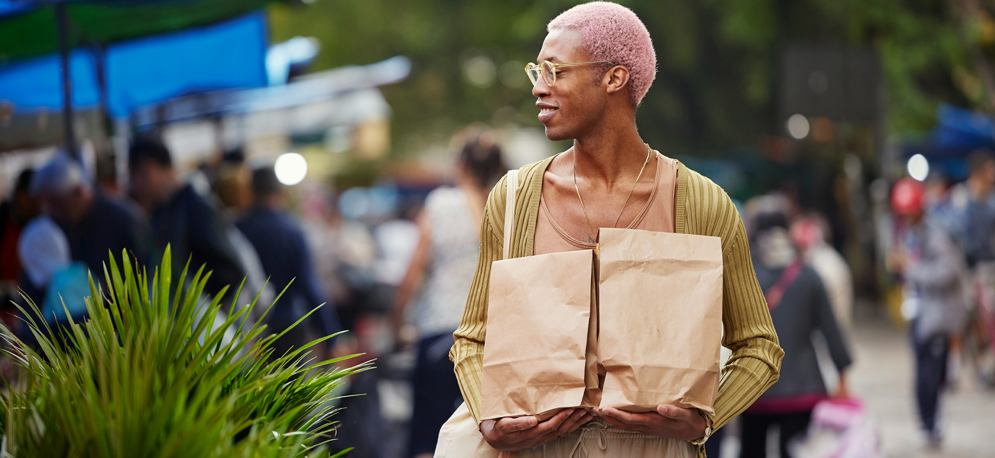 Eine Person mit pinken Haaren trägt zwei braune Papiertüten und lächelt. Sie steht auf einem belebten Markt, umgeben von Menschen und bunten Markisen. Die Atmosphäre ist lebhaft und freundlich.