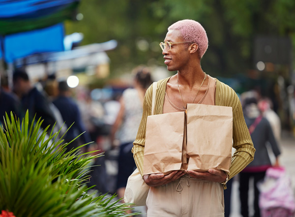 Eine Person mit pinken Haaren trägt zwei braune Papiertüten und lächelt. Sie steht auf einem belebten Markt, umgeben von Menschen und bunten Markisen. Die Atmosphäre ist lebhaft und freundlich.