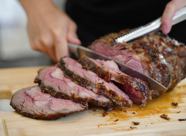 Eine Person schneidet ein großes Stück Fleisch mit einem Messer auf einem Schneidebrett. Das Fleisch ist perfekt gegart und zeigt eine saftige, rosa Innenseite. Die Hand hält das Messer, während die andere Hand das Fleisch stabilisiert.