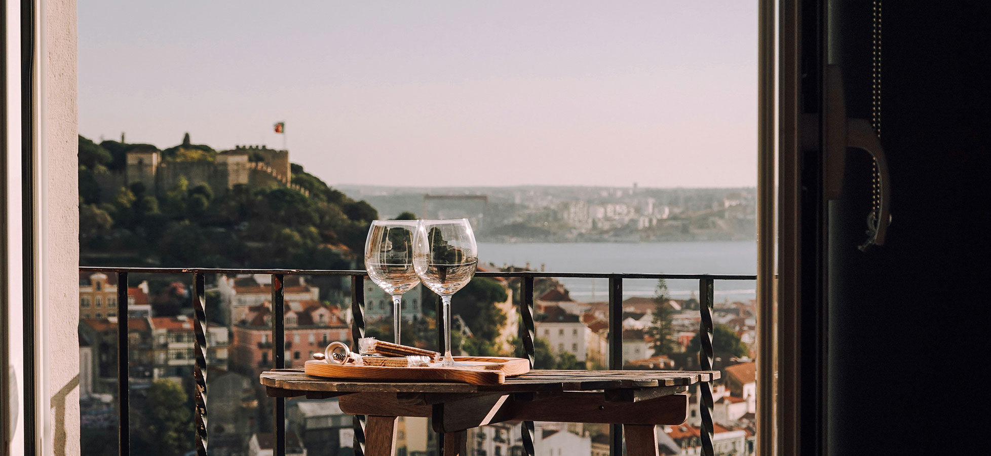 Zwei Weingläser stehen auf einem kleinen Tisch, der auf einem Balkon steht. Im Hintergrund sind sanfte Hügel und eine Stadtlandschaft mit Häusern sowie ein Wasserlauf zu sehen. Die Atmosphäre wirkt einladend und entspannend.