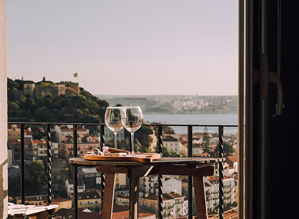 Zwei Weingläser stehen auf einem kleinen Tisch, der auf einem Balkon steht. Im Hintergrund sind sanfte Hügel und eine Stadtlandschaft mit Häusern sowie ein Wasserlauf zu sehen. Die Atmosphäre wirkt einladend und entspannend.