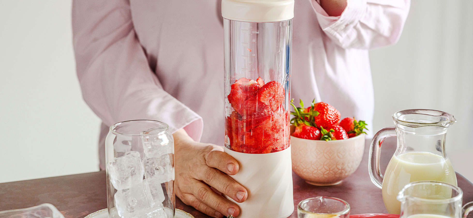 Eine Person hält einen Mixer, in dem frische Erdbeeren gemixt werden. Auf dem Tisch stehen Eiswürfel, ein Glas, eine Schüssel mit weiteren Erdbeeren und ein Krug Milch. Die Umgebung wirkt frisch und einladend.