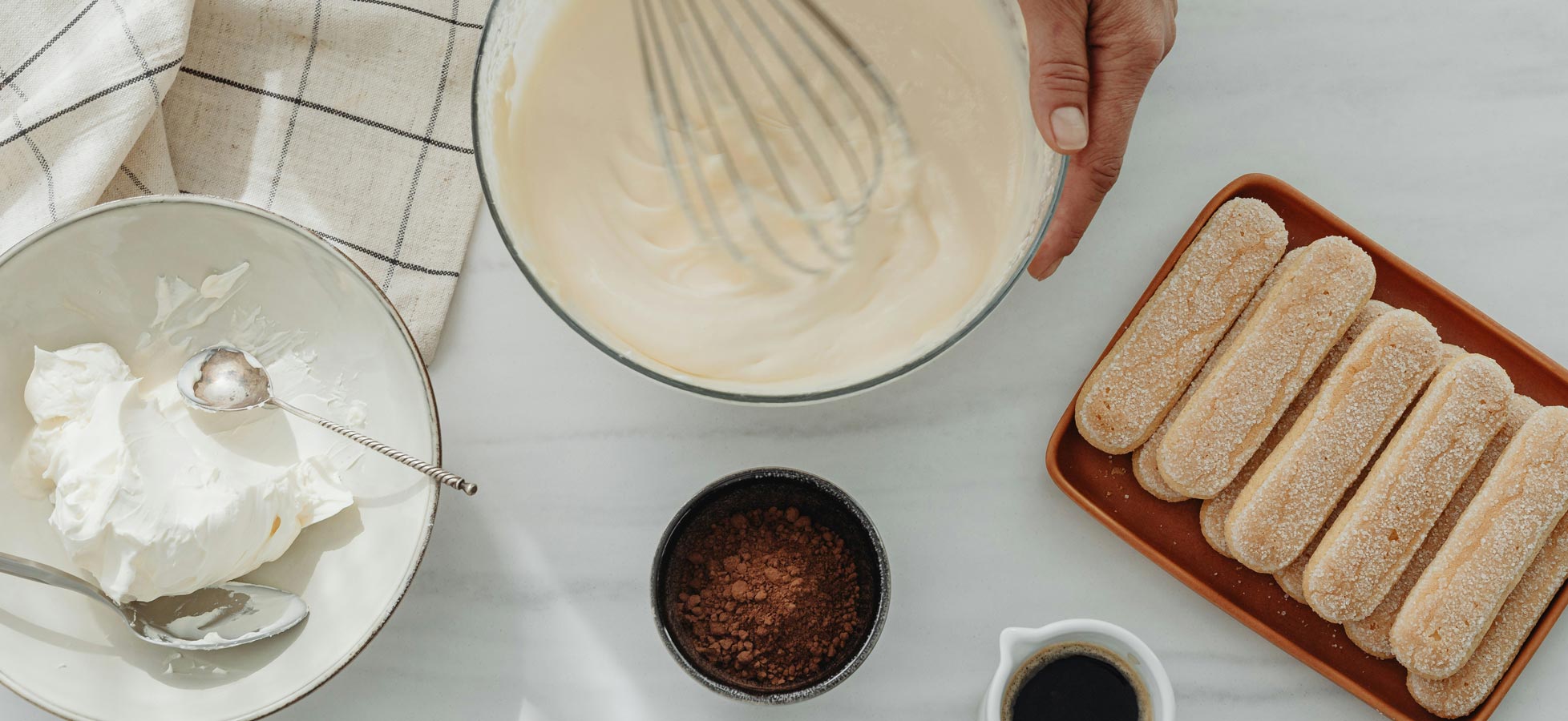 Eine Hand hält einen Schneebesen über eine Schüssel mit cremiger Füllung. Neben der Schüssel sind Löffel, ein Behälter mit Kakaopulver und eine Platte mit gezuckerten Löffelbiskuits angeordnet. Ein Geschirrtuch mit Karomuster liegt im Bild.
