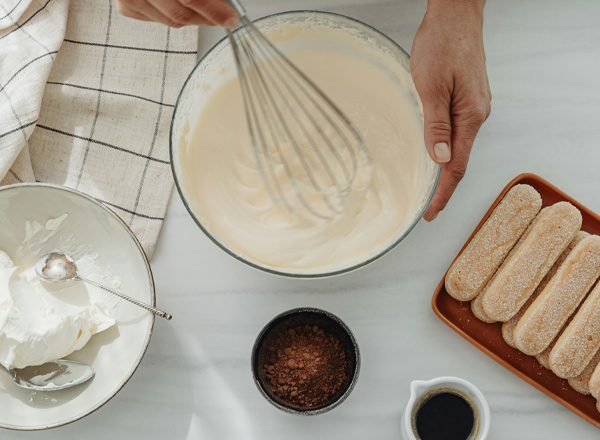 Eine Hand hält einen Schneebesen und rührt eine cremige Masse in einer Schüssel. Neben der Schüssel liegen Löffel mit Sahne, eine Schale mit Kakaopulver und ein Teller mit Löffelbiskuits. Der Hintergrund zeigt ein Küchentuch mit kariertem Muster.