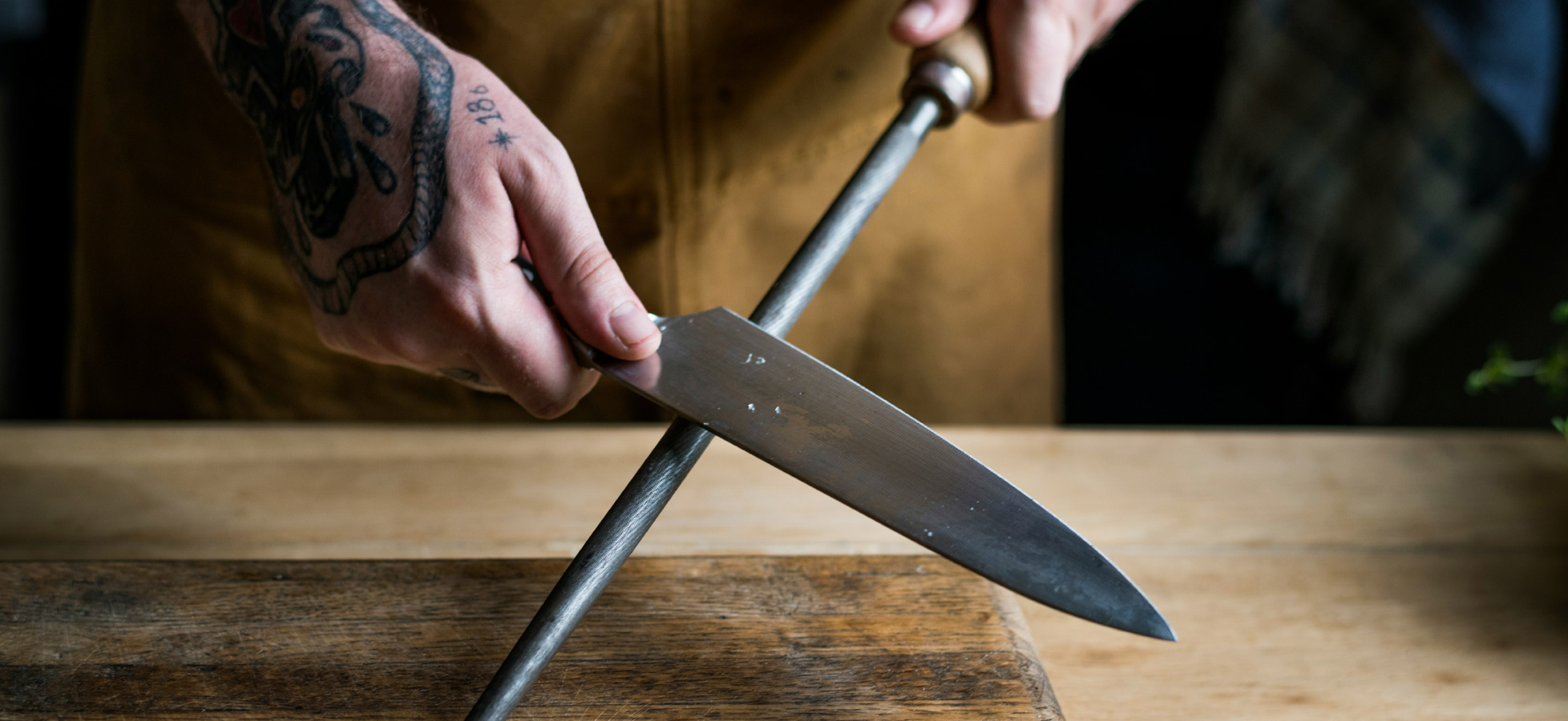 Ein Mensch mit tätowierten Händen schleift ein Küchenmesser mit einem Wetzstahl auf einem Holzbrett. Die Bewegung zeigt eine effektive Technik zur Schärfung des Messers, die die Zubereitung von Lebensmitteln erleichtert.