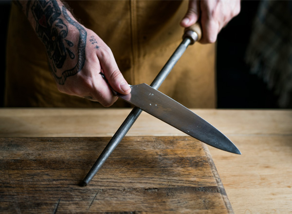 Ein Mensch mit tätowierten Händen schleift ein Küchenmesser mit einem Wetzstahl auf einem Holzbrett. Die Bewegung zeigt eine effektive Technik zur Schärfung des Messers, die die Zubereitung von Lebensmitteln erleichtert.