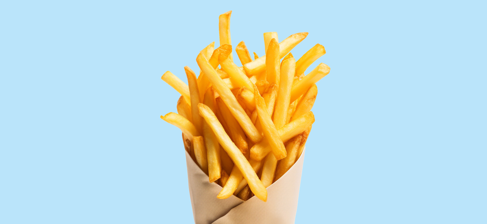 Ein Pappbecher mit knusprigen Pommes frites, die goldbraun und frisch zubereitet sind. Die Pommes stehen hoch aus dem Becher heraus und sind vor einem hellblauen Hintergrund platziert.