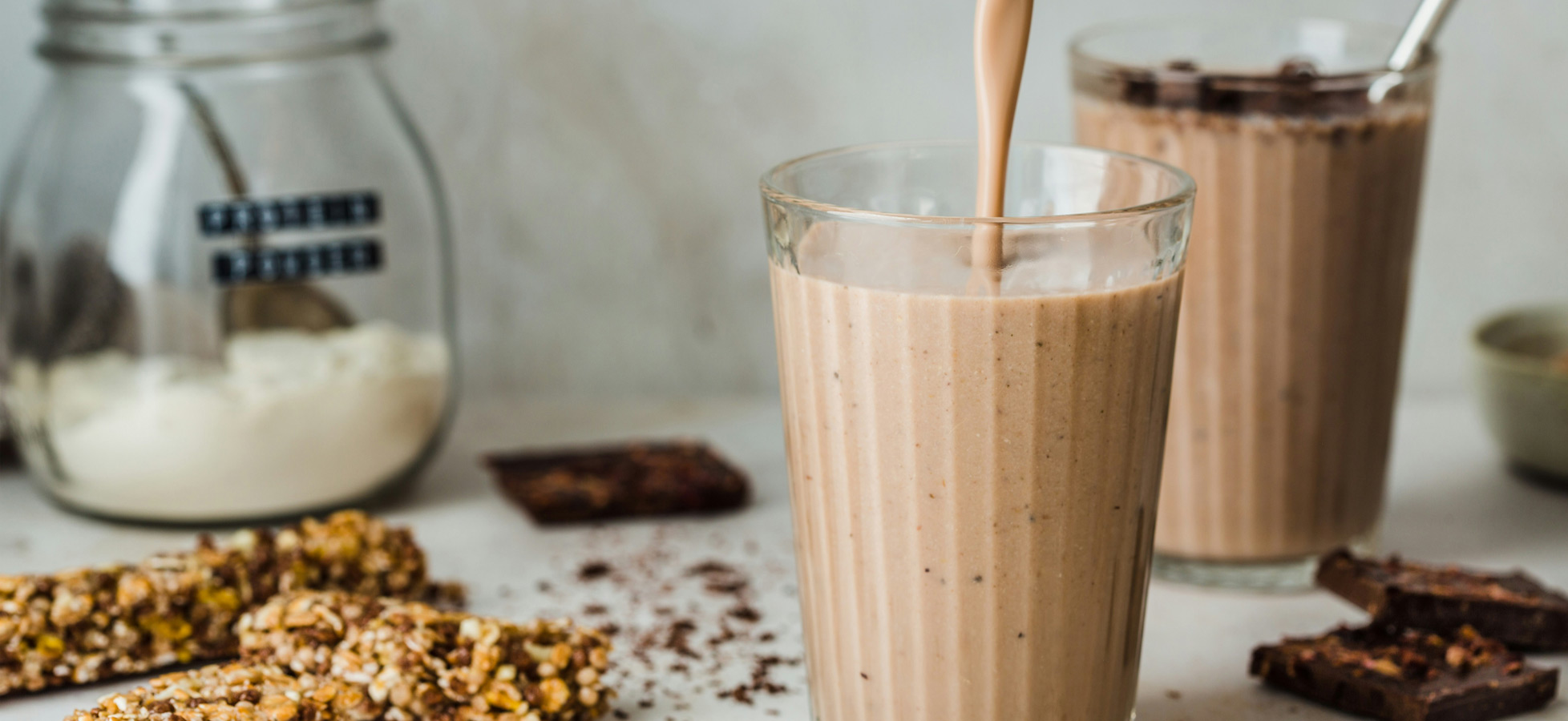 Ein Glas mit einem schokoladenfarbenen Smoothie wird gerade gefüllt. Im Hintergrund sind zwei weitere Gläser mit ähnlichem Inhalt und einige Snacks wie Müsliriegel und Schokolade zu sehen. Die Umgebung wirkt hell und gemütlich.