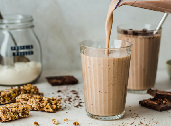Ein Glas mit einem schokoladenfarbenen Smoothie wird gerade gefüllt. Im Hintergrund sind zwei weitere Gläser mit ähnlichem Inhalt und einige Snacks wie Müsliriegel und Schokolade zu sehen. Die Umgebung wirkt hell und gemütlich.