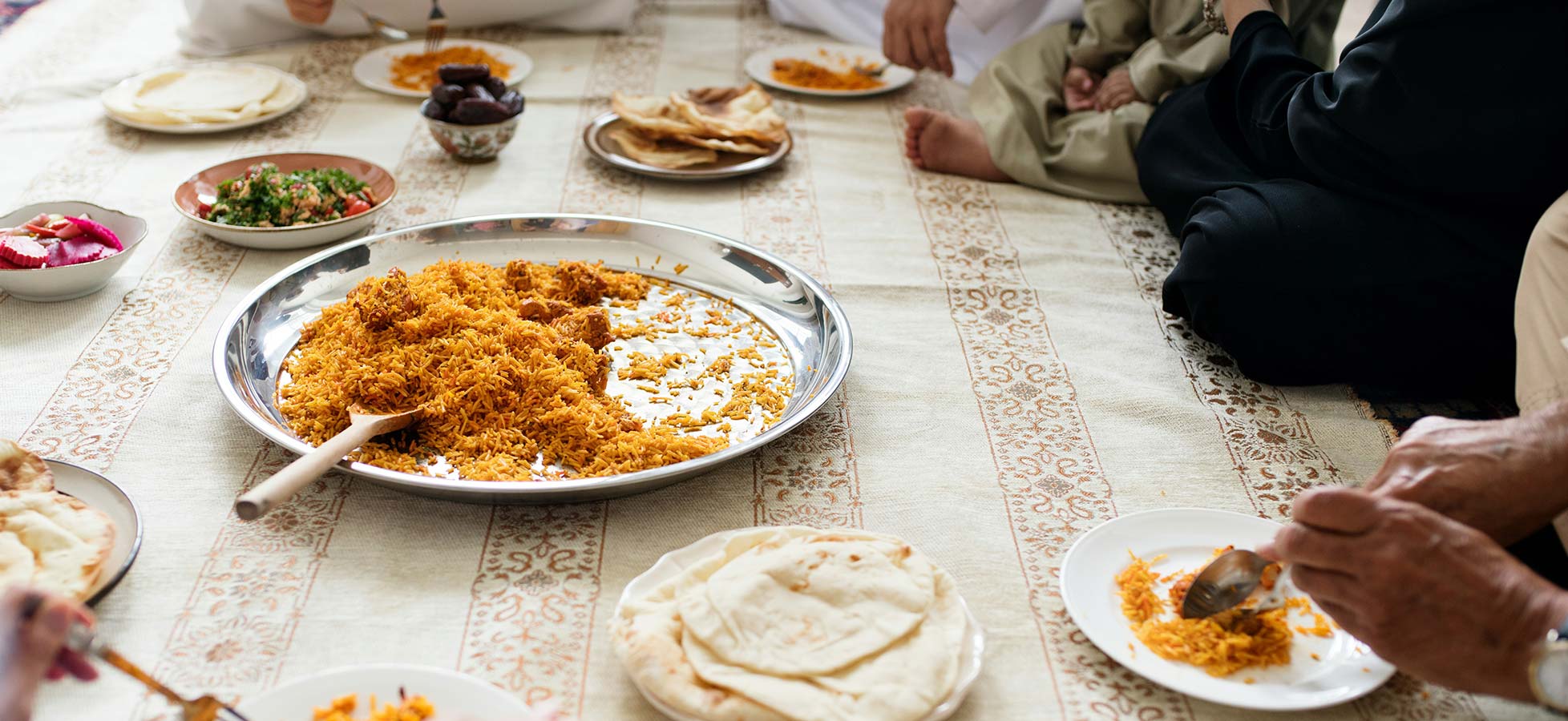 Eine große Platten mit Reis und verschiedenen Beilagen liegt auf einem Tisch. Um den Tisch herum sitzen Personen, die essen. Auf dem Tisch sind auch Fladenbrot und Salat angerichtet. Die Atmosphäre vermittelt Gemeinschaft und ein festliches Essen.