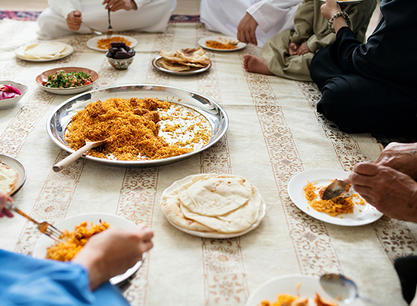 Eine Gruppe von Personen sitzt um einen Tisch und genießt eine Mahlzeit. Im Mittelpunkt steht ein großer Teller mit Biryani, umgeben von kleinen Tellern mit flachen Broten und Salat. Einige Personen verwenden Besteck, um ihr Essen zu servieren.